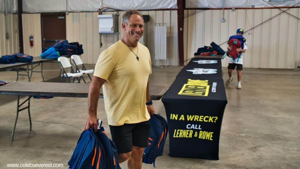 smiling Glen Lerner, wearing a yellow shirt and black shorts, carries blue and orange backpacks inside a spacious hall with tables, chairs, and a "Lerner & Rowe" branded tablecloth advertising legal services.