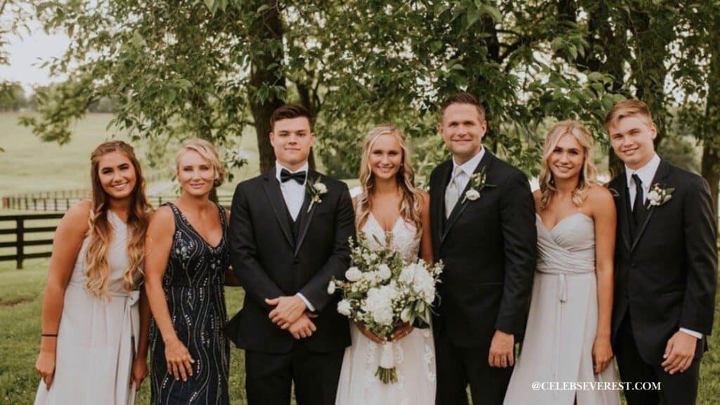 A wedding group photo featuring Kyle Idleman, dressed in a suit, standing next to a woman in a white wedding dress holding a bouquet of flowers. They are surrounded by other formally dressed people, including a man in a tuxedo and women in elegant dresses, against a backdrop of trees and a grassy field.